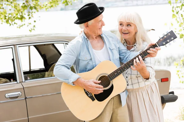 Marido y mujer mayores mirándose y tocando la guitarra contra el coche beige - foto de stock