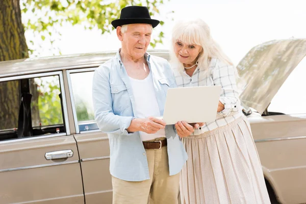 Feliz pareja de ancianos mirando a la computadora portátil contra coche vintage - foto de stock