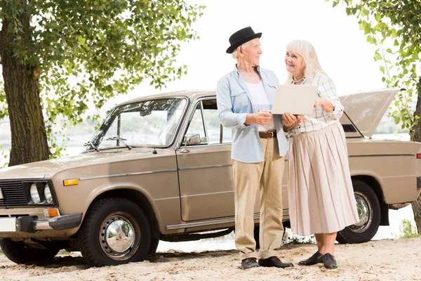 Hermosa pareja de ancianos utilizando el ordenador portátil y mirándose unos a otros contra el coche vintage beige - foto de stock