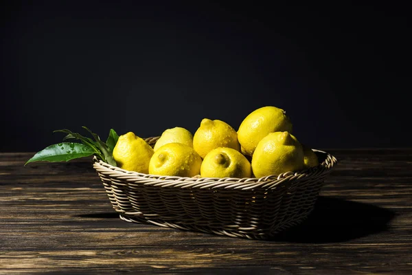 Limones amarillos con hojas en canasta de mimbre sobre mesa de madera con fondo negro - foto de stock