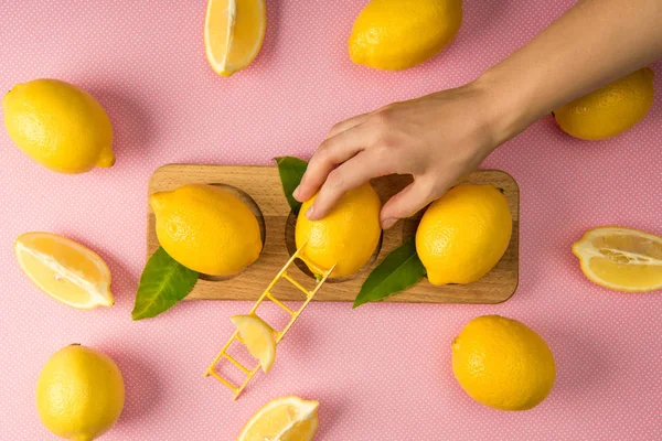 Vista recortada de la mujer tomando limón con pequeña escalera de madera sobre fondo rosa con limones frescos - foto de stock