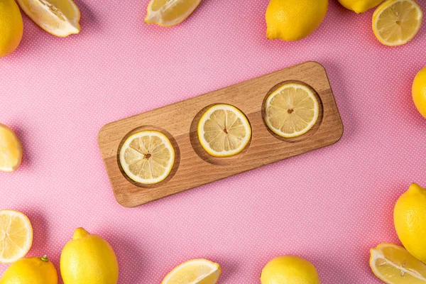 Вид сверху на свежие ломтики лимона на деревянной доске с лимонами вокруг на розовом фоне — стоковое фото