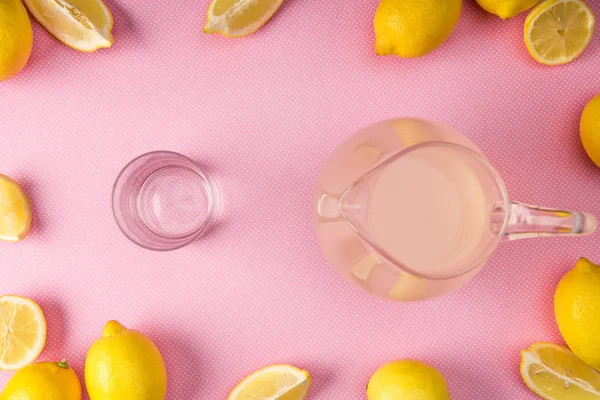 Вид сверху на стекло и кувшин со свежим соком на розовом фоне с лимонами — стоковое фото