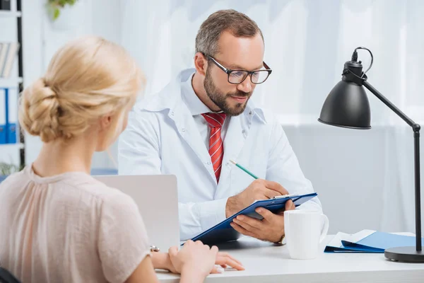 Physiothérapeute en manteau blanc prenant des notes dans un bloc-notes avec une patiente à proximité lors d'un rendez-vous à la clinique — Photo de stock