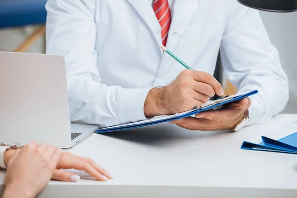 Partial view of physiotherapist in white coat making notes in notepad with female patient near by during appointment in clinic — Stock Photo