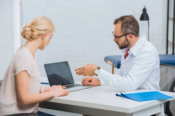 Фізіотерапевт у білому пальто розмовляє з пацієнткою на робочому місці з ноутбуком в лікарні — стокове фото