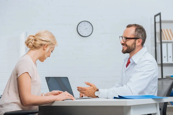 Fisioterapeuta con bata blanca conversando con paciente femenina en el lugar de trabajo con portátil en el hospital - foto de stock