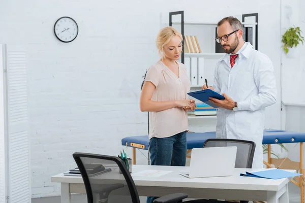 Portrait of chiropractic in white coat with notepad and female patient having conversation in clinic — Stock Photo