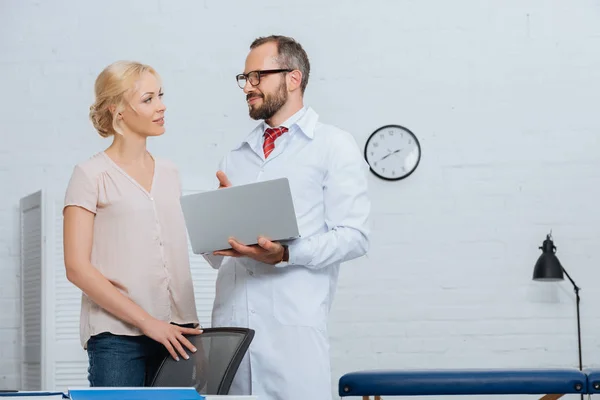 Fisioterapista in camice bianco con laptop e paziente donna che si guarda in ospedale — Foto stock
