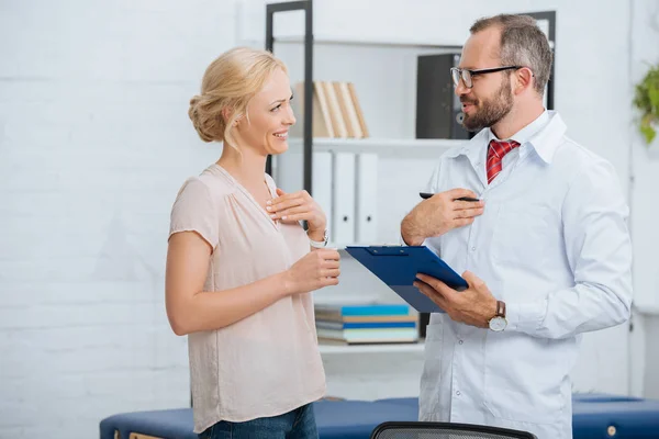 Side view of chiropractic in white coat with notepad and smiling female patient in clinic — Stock Photo