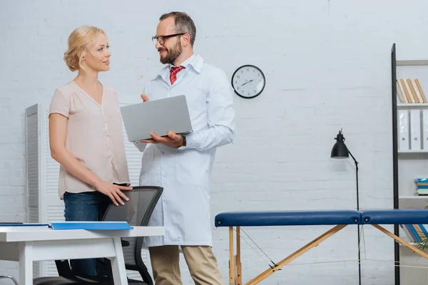 Physiotherapist in white coat with laptop and female patient looking at each other in hospital — Stock Photo