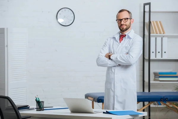 Quiroprático masculino em casaco branco e óculos com braços cruzados de pé no local de trabalho com laptop no hospital — Fotografia de Stock