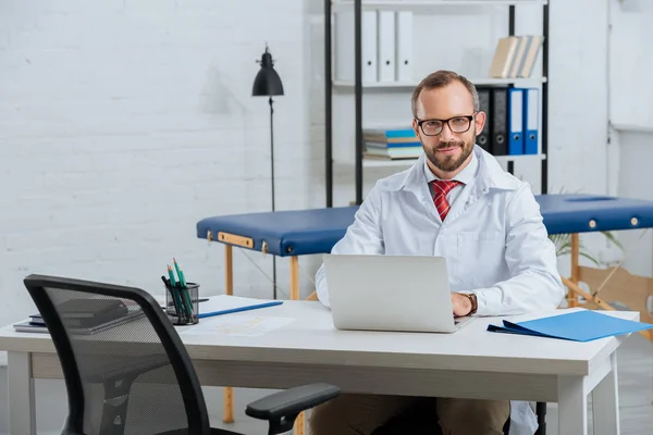 Retrato de quiroprático masculino sorridente em casaco branco no local de trabalho com laptop no hospital — Fotografia de Stock