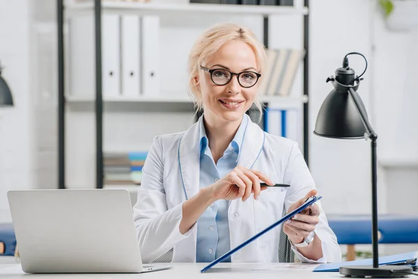 Porträt eines lächelnden Chiropraktikers mit Brille und weißem Mantel, der auf Notizblock am Arbeitsplatz in der Klinik zeigt — Stockfoto