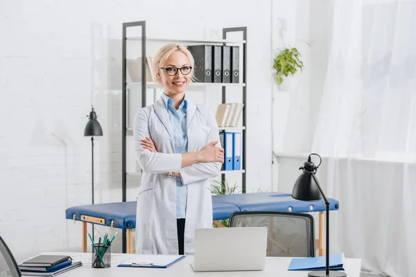 Portrait of smiling chiropractor in eyeglasses and white coat standing at workplace in clinic — Stock Photo