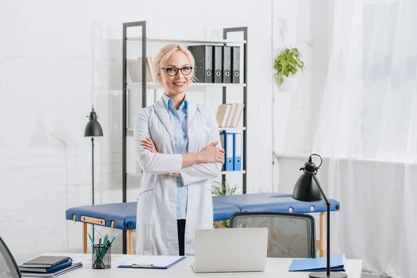 Retrato de quiropráctico sonriente en gafas y bata blanca de pie en el lugar de trabajo en la clínica - foto de stock