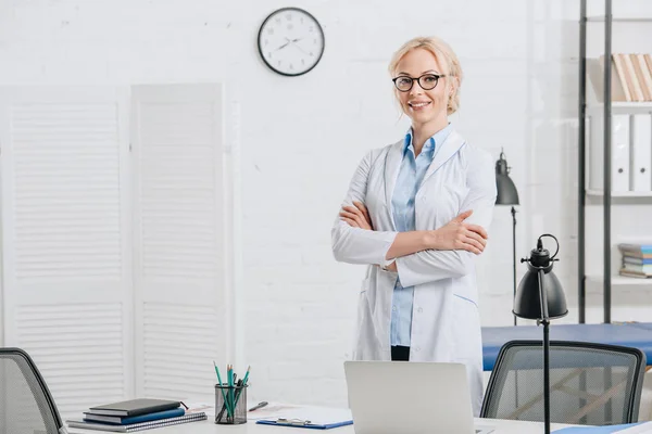 Retrato de quiropráctico sonriente en gafas y bata blanca de pie en el lugar de trabajo en la clínica - foto de stock