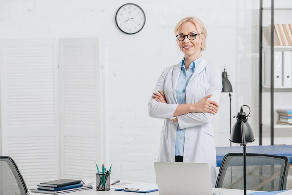 Ritratto di chiropratico sorridente in occhiali e cappotto bianco in piedi sul posto di lavoro in clinica — Foto stock