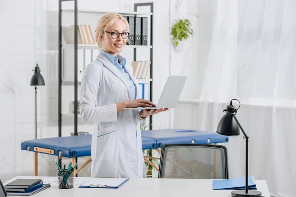 Vue latérale du chiropraticien souriant en lunettes et manteau blanc avec ordinateur portable en clinique — Photo de stock