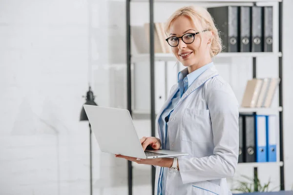 Seitenansicht eines lächelnden Chiropraktikers mit Brille und weißem Mantel mit Laptop in der Klinik — Stockfoto