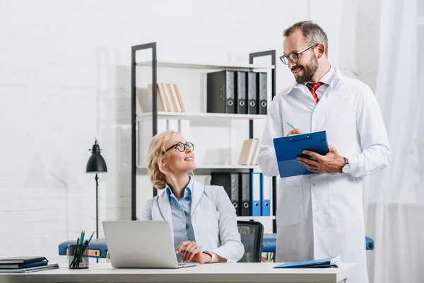 Fisioterapeutas sonrientes con batas blancas mirándose en el lugar de trabajo con portátil en la clínica - foto de stock