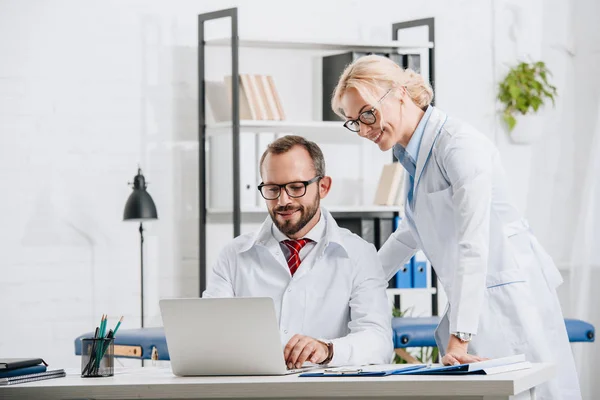 Smiling physiotherapists in white coats using laptop together in clinic — Stock Photo