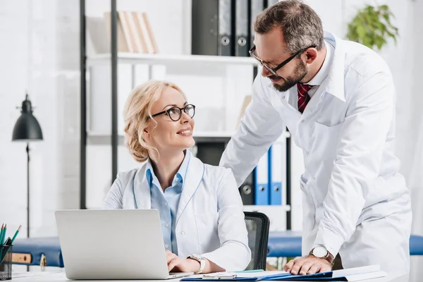 Fisioterapeutas sonrientes con batas blancas mirándose en el lugar de trabajo con portátil en la clínica - foto de stock