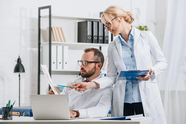 Portrait of physiotherapists in white coats discussing picture at workplace with laptop in clinic — Stock Photo