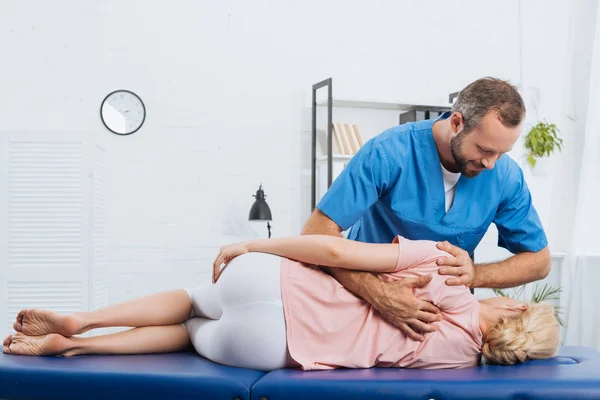 Quiropráctico sonriente masajeando la espalda del paciente que yace en la mesa de masaje en el hospital - foto de stock