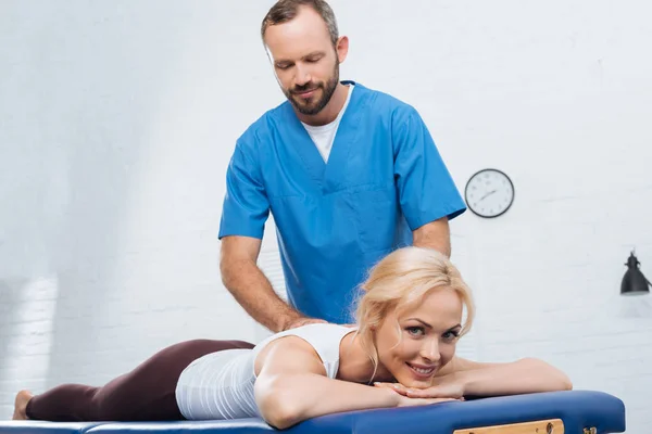Masajista haciendo masaje a mujer sonriente en mesa de masaje en clínica - foto de stock