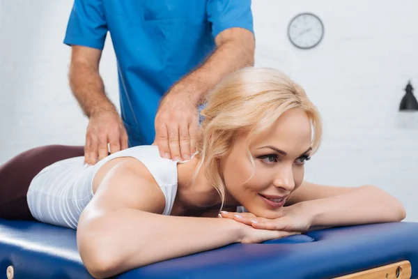 Vista parziale del massaggiatore che fa massaggi alla donna sorridente sul tavolo da massaggio in clinica — Foto stock