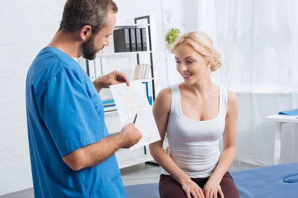 Physiothérapeute montrant schéma du corps humain à la femme sur la table de massage à l'hôpital — Photo de stock