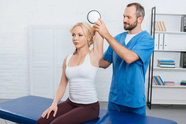 Chiropraticien souriant étirant le bras des femmes sur la table de massage à la clinique — Photo de stock