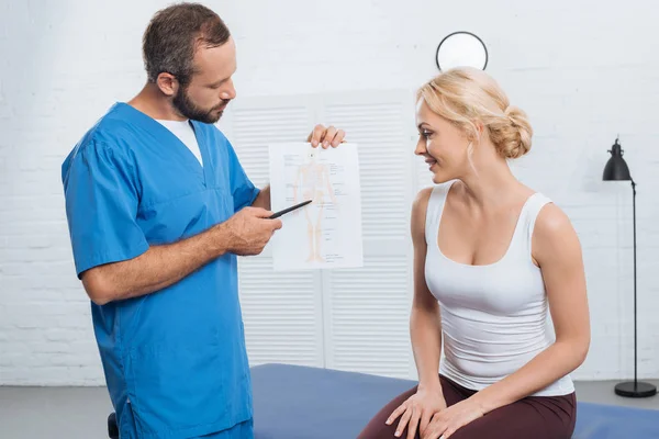 Physiotherapist showing human body scheme to smiling woman on massage table in hospital — Stock Photo