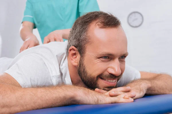 Частичный взгляд массажиста, делающего массаж улыбающемуся пациенту на массажном столе в клинике — стоковое фото