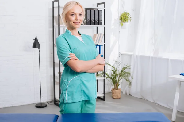 Portrait de masseuse souriante aux bras croisés debout à la table de massage à la clinique — Photo de stock