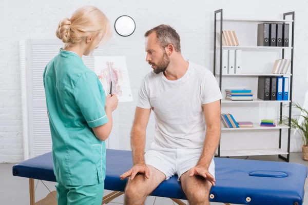 Chiropraktik zeigt männlichen Patienten während eines Termins in der Klinik das menschliche Körperbild auf dem Massagetisch — Stockfoto
