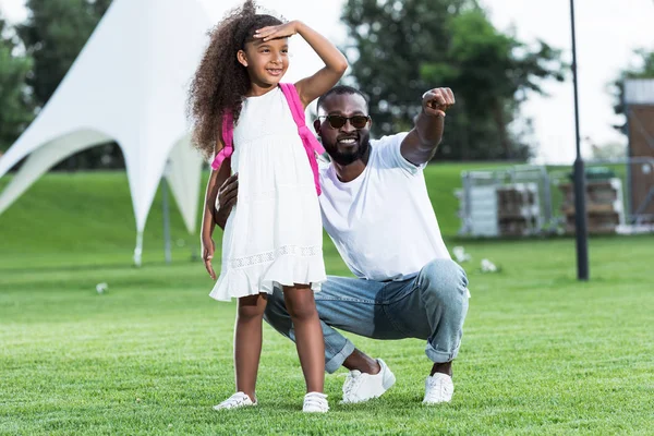 Sorrindo afro-americano pai apontando em algo para filha com saco escolar no parque — Fotografia de Stock