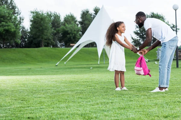 African american daughter giving school bag to father in park — Stock Photo