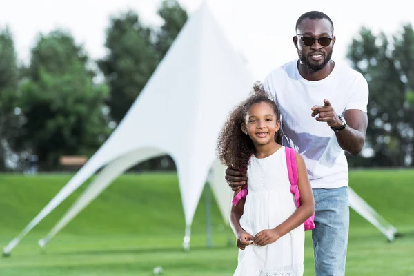 African american father pointing on camera to adorable daughter with school bag in park — Stock Photo