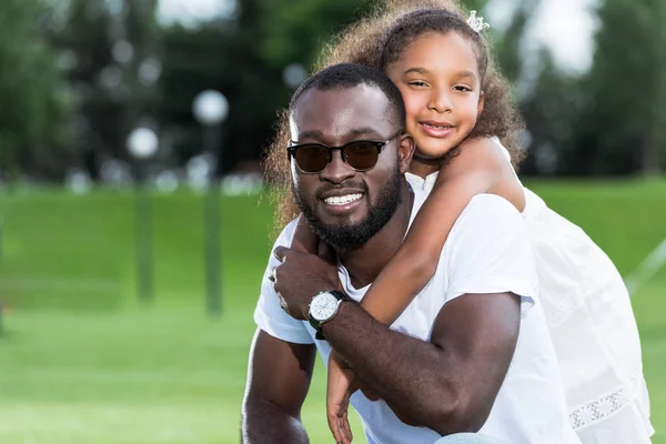 Hija afroamericana abrazando sonriente padre de vuelta en el parque - foto de stock