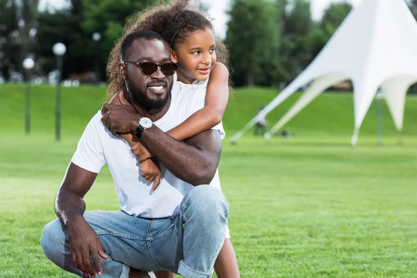 Afro-americana filha abraçando pai de volta no parque e eles olhando para longe — Fotografia de Stock