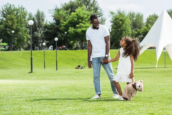 Afro-americanos padre e hija con osito de peluche cogidos de la mano y caminando en parque - foto de stock