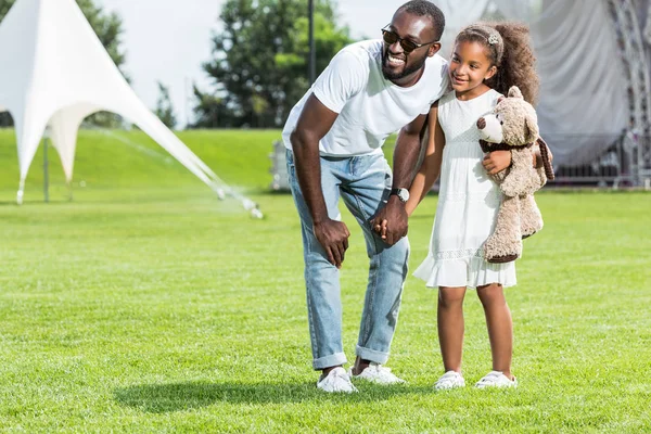 Padre e hija afroamericanos tomados de la mano en el parque y mirando hacia otro lado - foto de stock