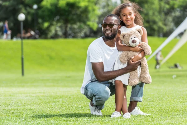Souriant afro-américain père accroupi et câlin fille avec ours en peluche dans le parc — Photo de stock