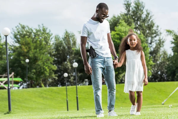 African american cop with gun in holster and daughter holding hands and walking at amusement park — Stock Photo