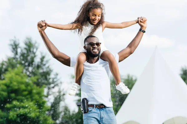 Счастливый африканский офицер полиции Америки с пистолетом, держащий дочь на плечах в парке развлечений — стоковое фото