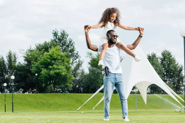 African american police officer with gun holding daughter on shoulders in amusement park — Stock Photo