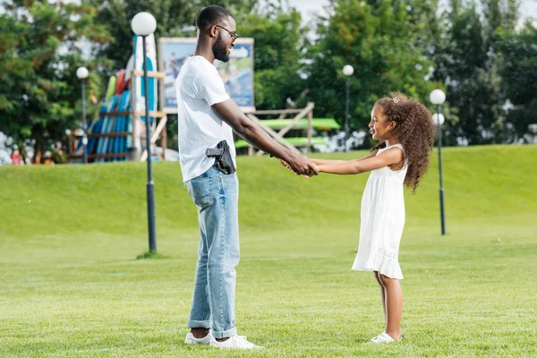 African american policeman with gun and daughter holding hands and spinning around in amusement park — Stock Photo
