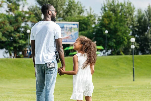 Vista posteriore di agente di polizia afroamericano e figlia che si tiene per mano e cammina nel parco divertimenti — Foto stock